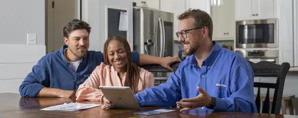 Three People Sit At A Kitchen Table, Reviewing Documents And A Tablet. The Man In The Blue Shirt Gestures At The Tablet, While The Other Two Listen Attentively With A Slight Smile.